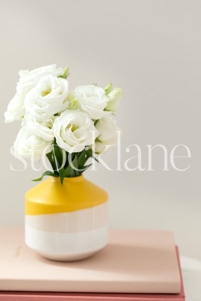 Vertical stock photo of bright yellow flower vase on pink notebooks