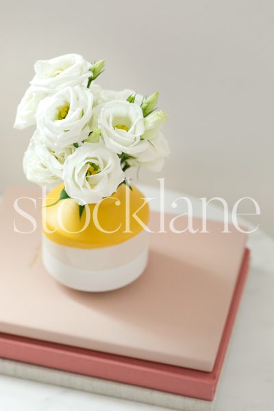 Vertical stock photo of bright yellow flower vase on pink notebooks