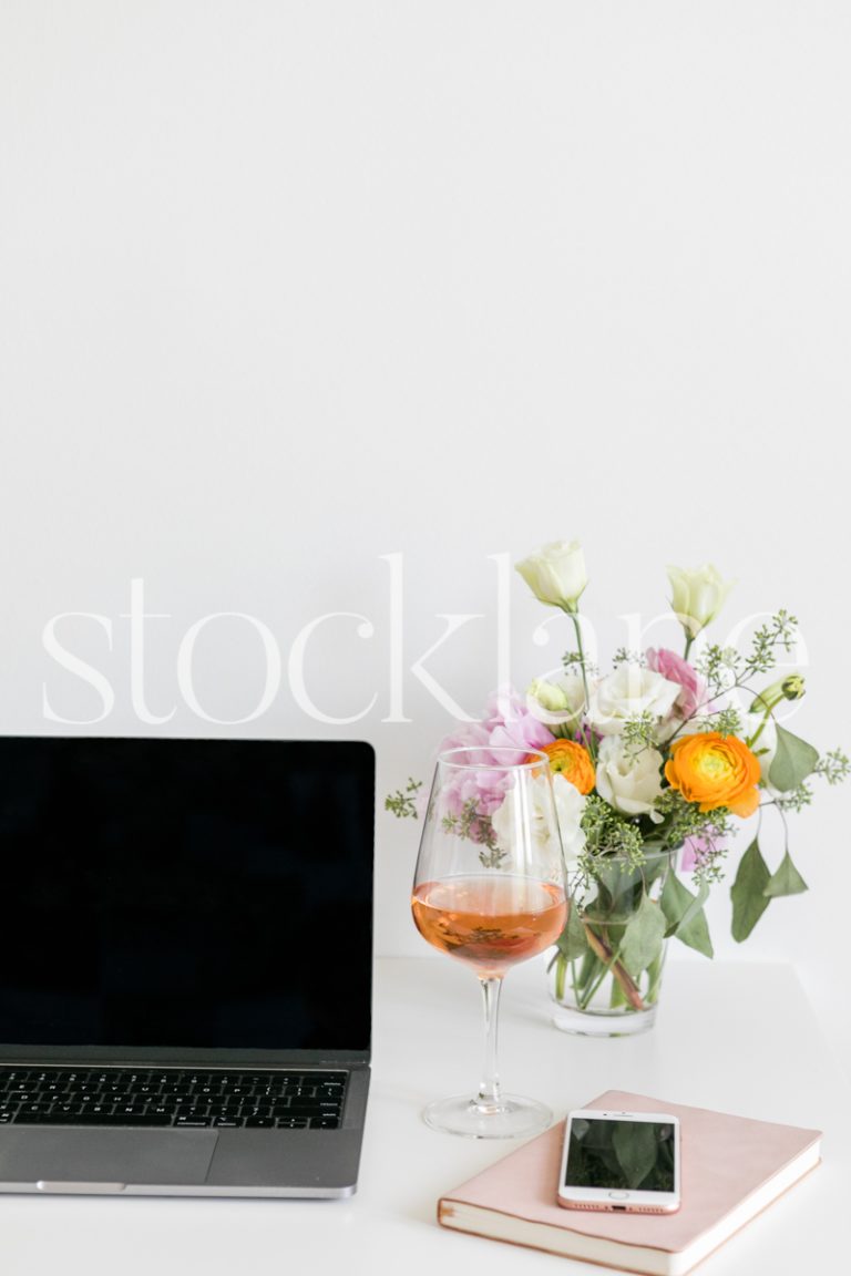 Vertical lifestyle stock photo of a feminine desktop with a computer, flowers and wine.