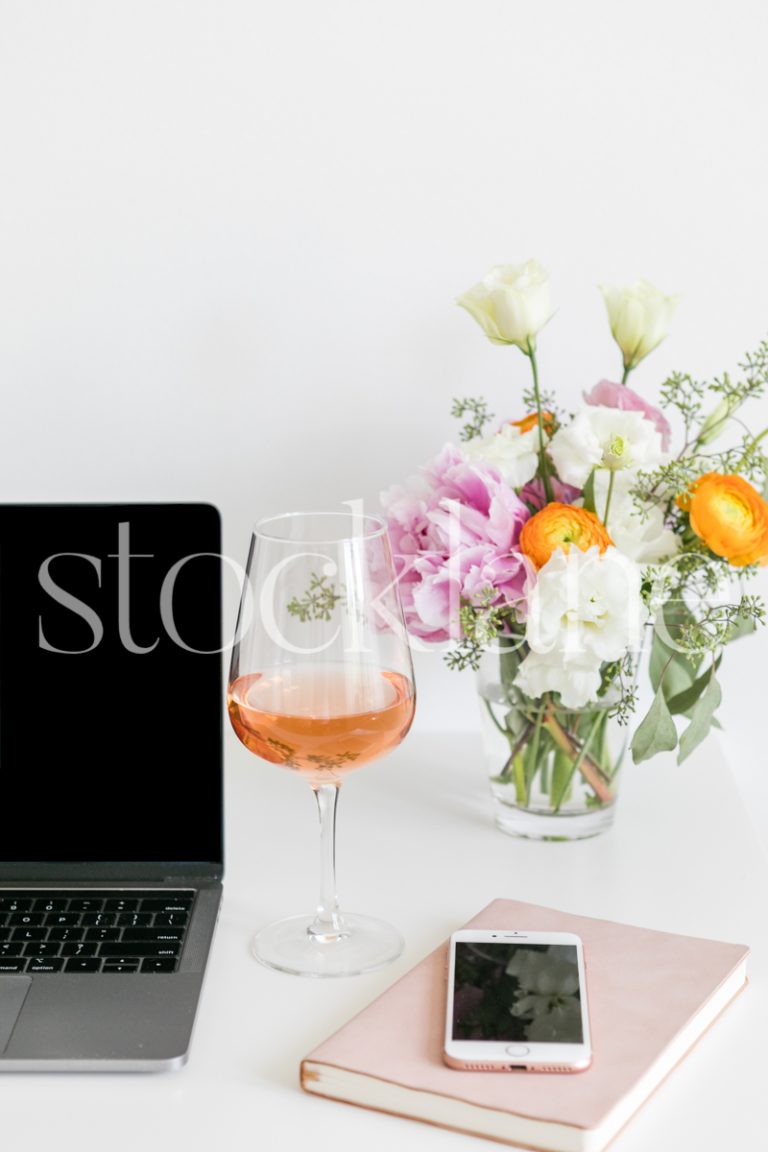 Vertical lifestyle stock photo of a feminine desktop with a computer, flowers and wine.