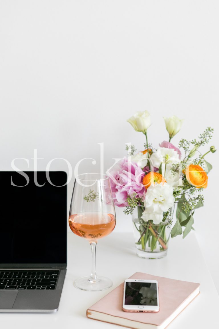 Vertical lifestyle stock photo of a feminine desktop with a computer, flowers and wine.