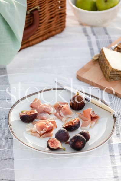 Vertical stock photo of plate with figs and prosciutto in beach picnic setting.