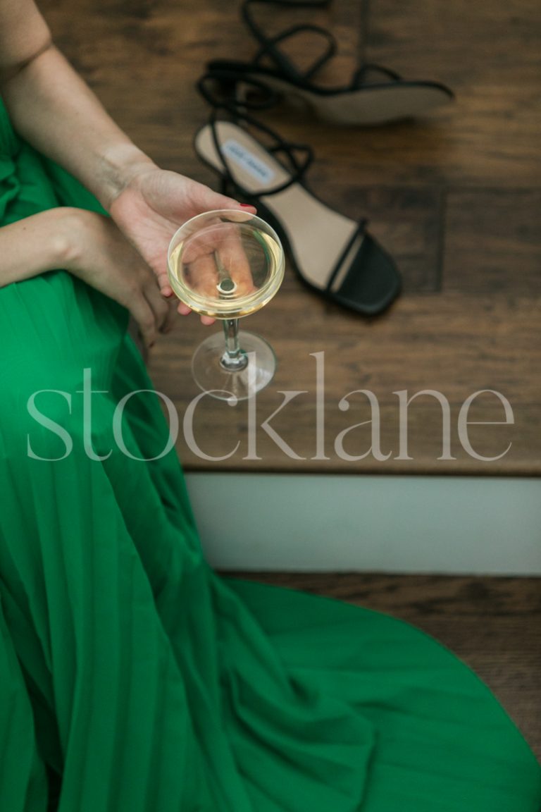 Vertical stock photo of a woman in a green dress sitting on stairs, holding a glass of champagne.