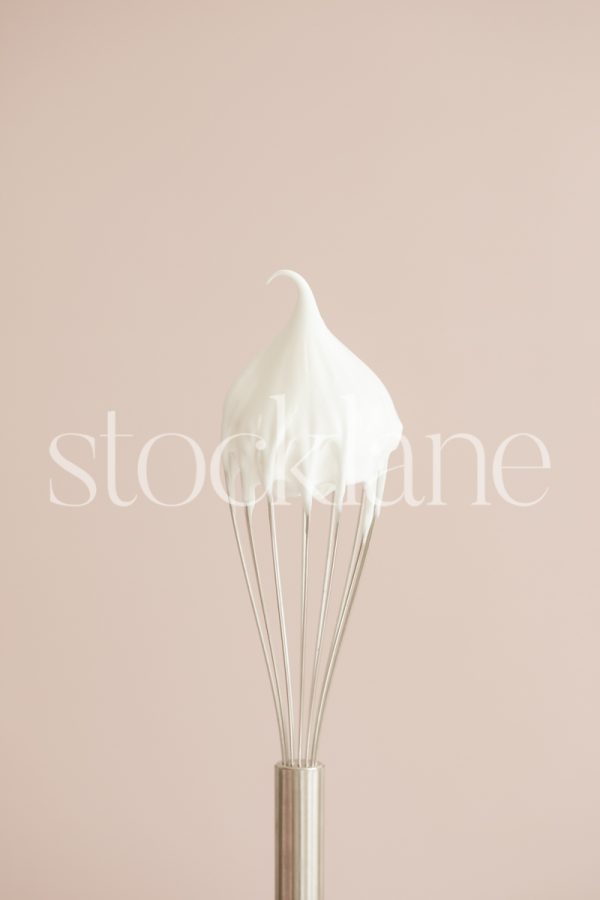 Vertical stock photo of a whisk with cream on a pink background