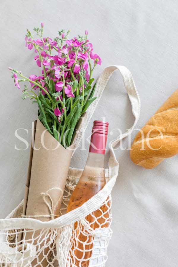 Vertical stock photo of a market bag with a flower bouquet, wine and bread.