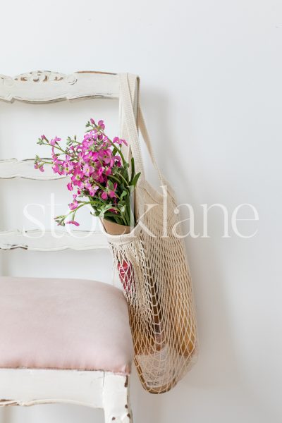 Vertical stock photo of a knit market bag with flowers and wine, hanging from a chair.