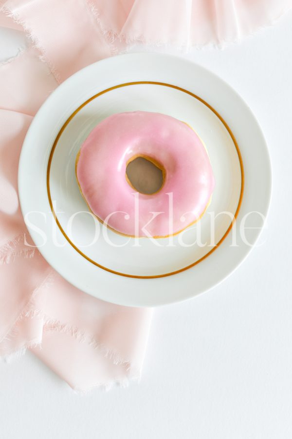 Vertical stock photo of a pink donut on a plate with a pink and white background.