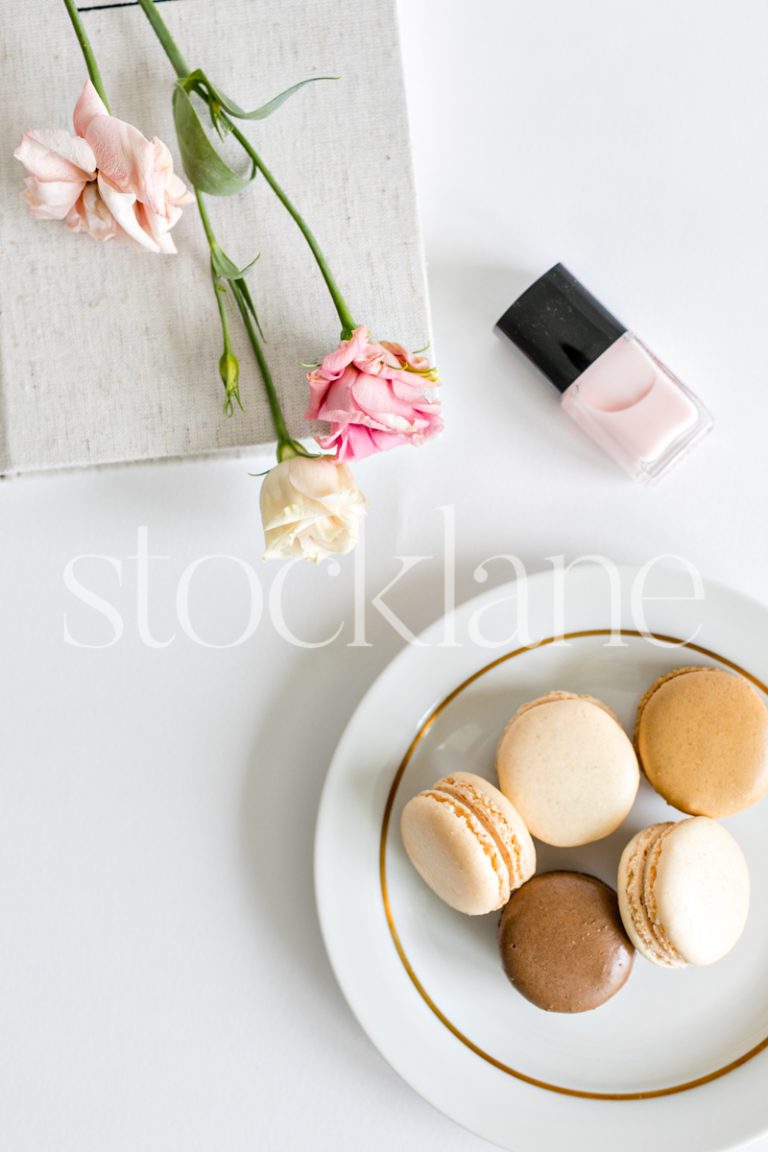 Vertical stock photo of a woman's desktop with flowers, nail polish and macarons.