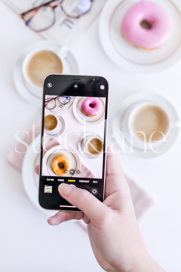 Vertical stock photo of a woman taking a phone picture of coffee and donuts.