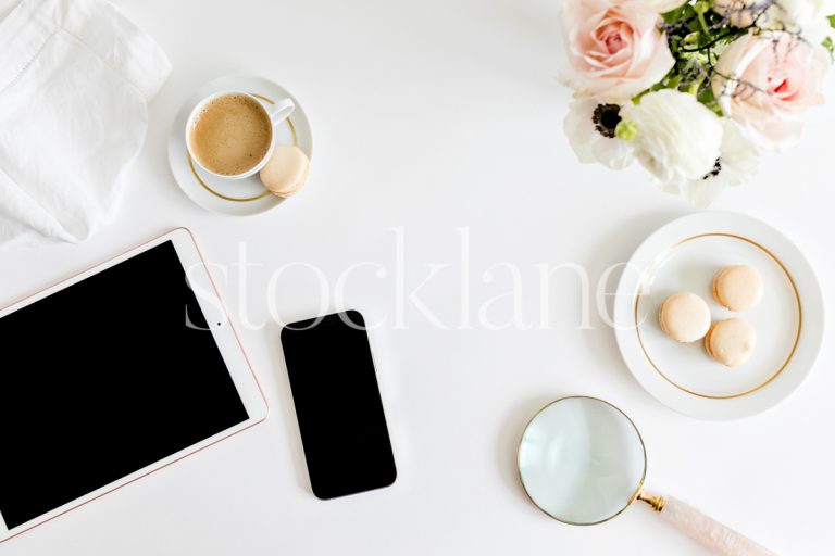 Horizontal photo of a woman's desktop with a tablet, phone, flowers, coffee and macarons.