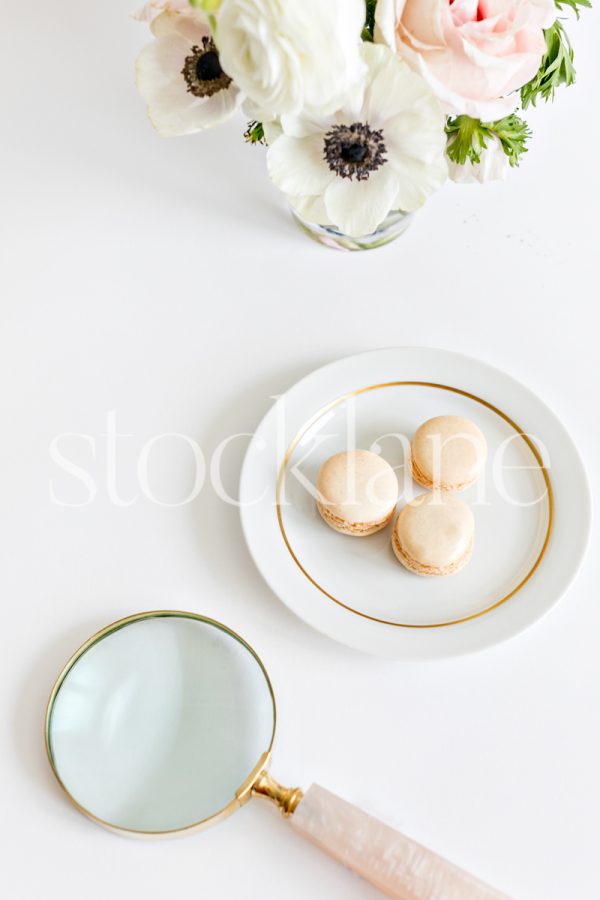 Vertical stock photo of a plate of vanilla macarons, a pink magnifying glass and flowers.