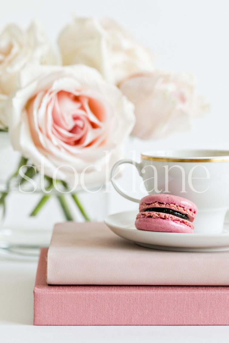 Vertical stock photo of a coffee cup on top of pink books, with a pink macaron and pink roses in the background.