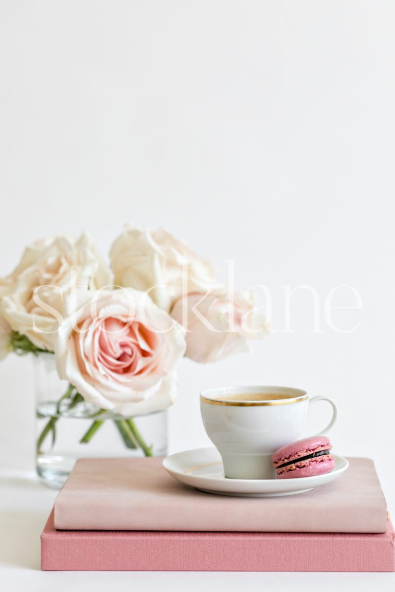 Vertical stock photo of a coffee cup on top of pink books, with a pink macaron and pink roses in the background.