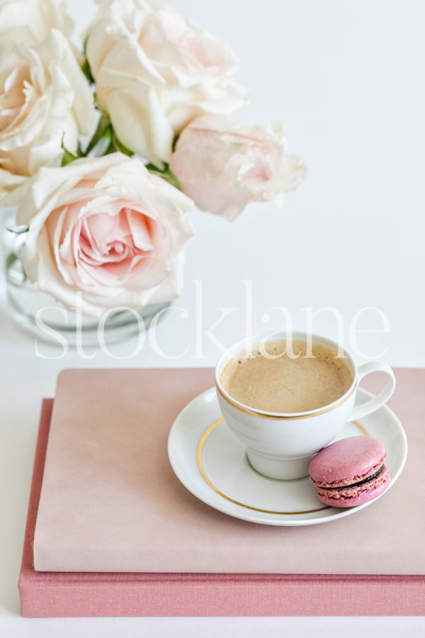 Vertical stock photo of a coffee cup on top of pink books, with a pink macaron and pink roses in the background.