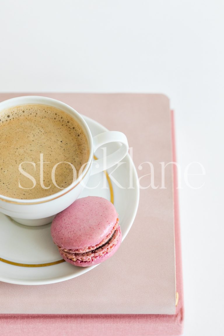 Vertical stock photo of a coffee cup on top of pink books, with a pink macaron.