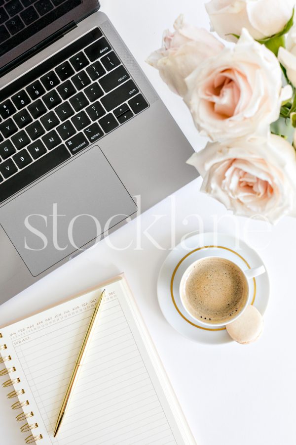 Vertical stock photo of a woman's desktop with a laptop, coffee, a notebook and pink roses.