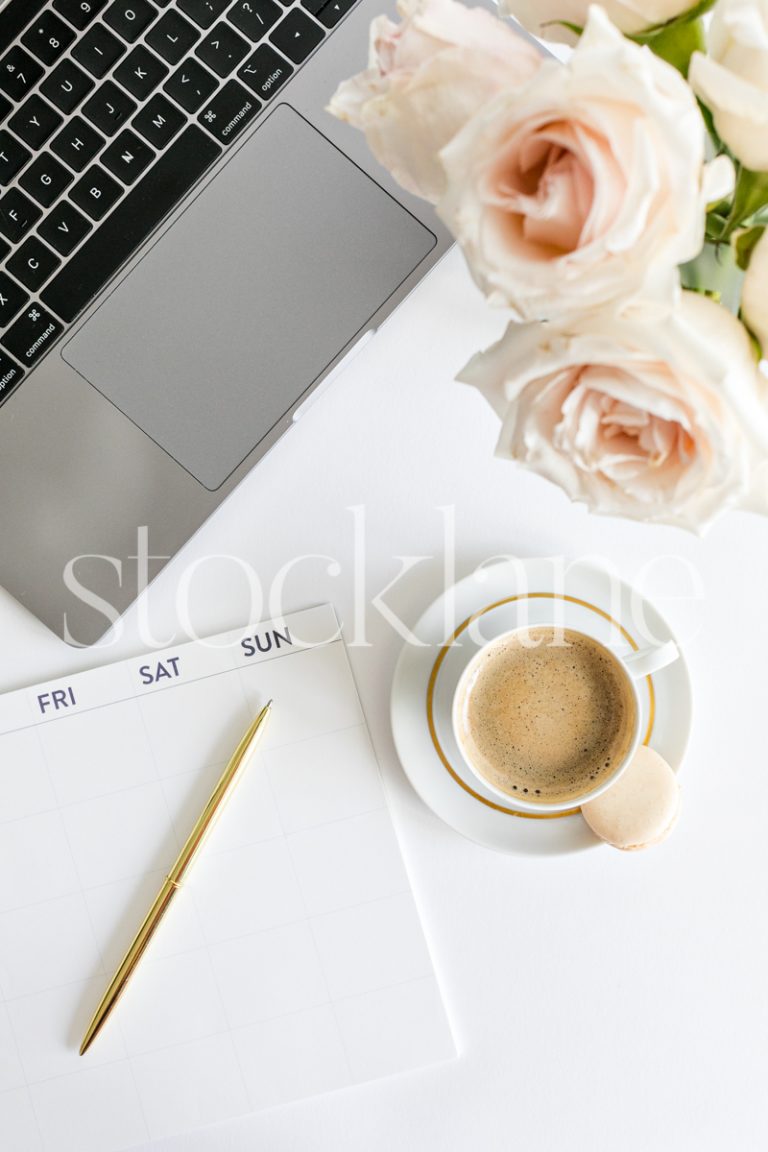 Vertical stock photo of a woman's desktop with a laptop, coffee, a calendar and pink roses.