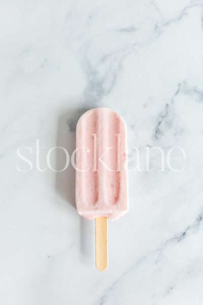 Vertical stock photo of a strawberry popsicle on a marble countertop.