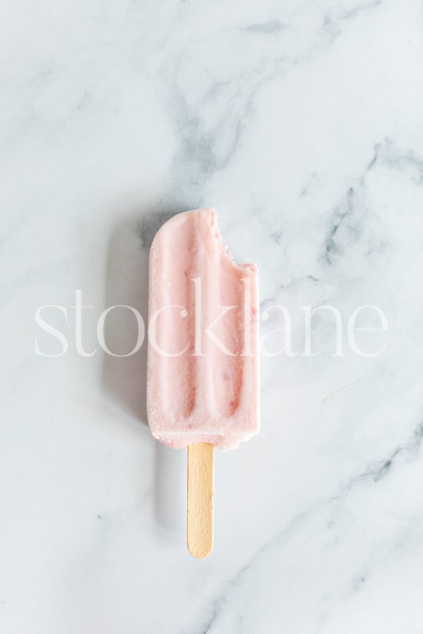 Vertical stock photo of a strawberry popsicle on a marble countertop.