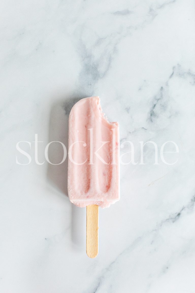 Vertical stock photo of a strawberry popsicle on a marble countertop.