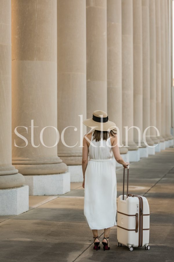 Vertical stock photo of a woman in a white dress with a suitcase in front of a classic building.