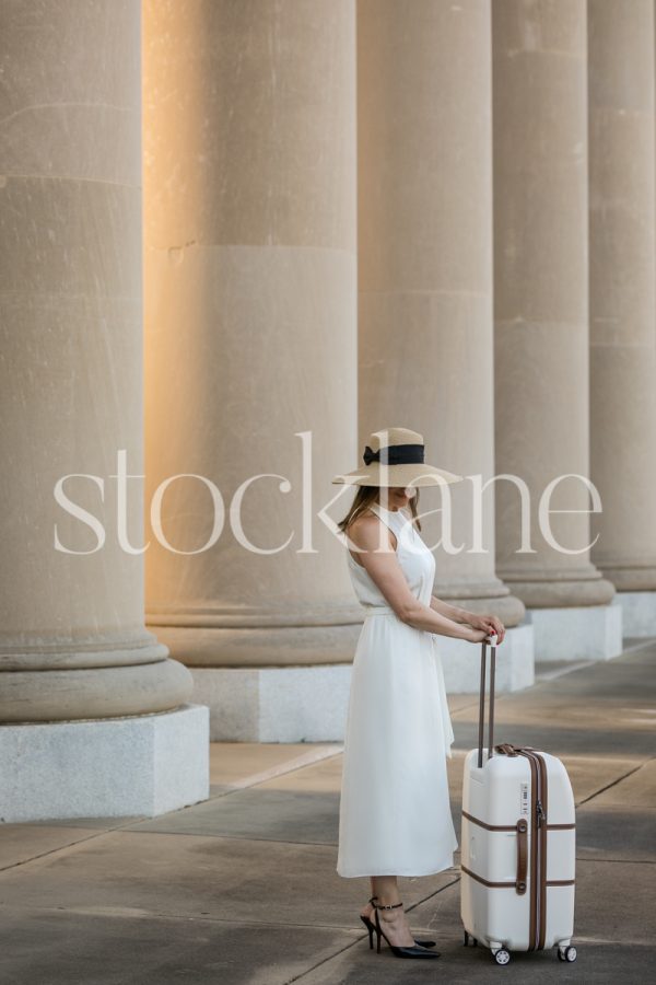 Vertical stock photo of a woman in a white dress with a suitcase in front of a classic building.