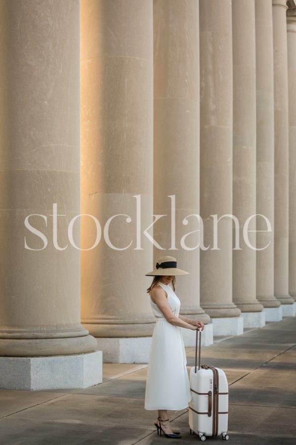 Vertical stock photo of a woman in a white dress with a suitcase in front of a classic building.