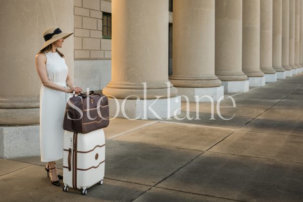 Horizontal stock photo of a woman in a white dress with a suitcase in front of a classic building.