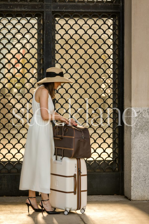 Vertical stock photo of a woman in a white dress with a suitcase in front of an ornate door with sunlight coming through.