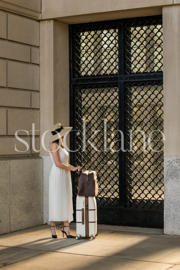 Vertical stock photo of a woman in a white dress with a suitcase in front of an ornate door with sunlight coming through.