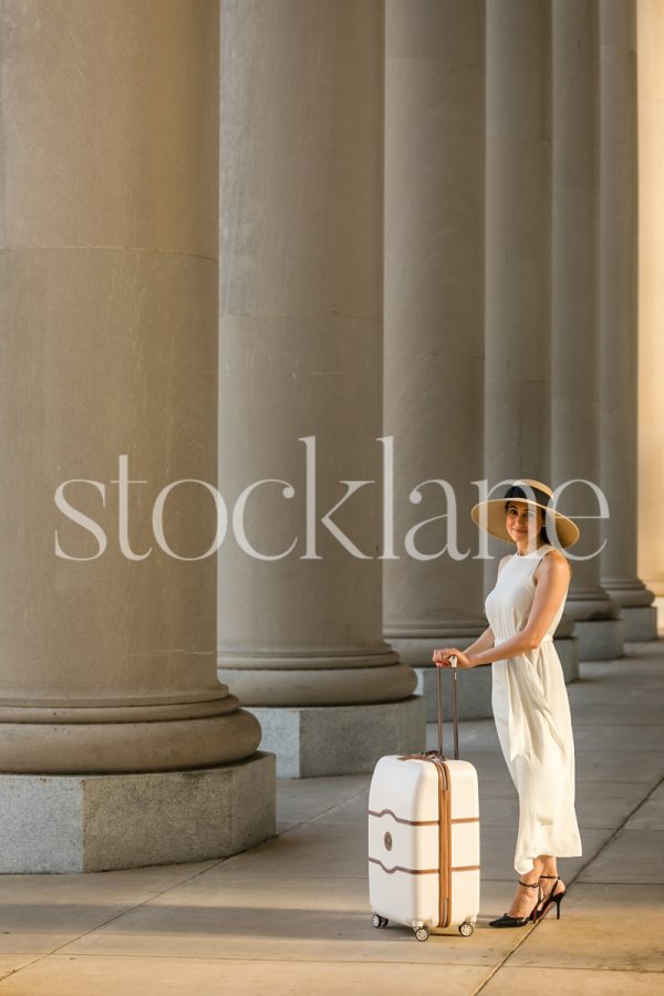 Vertical stock photo of a woman holding a suitcase in front of a classic building with columns.