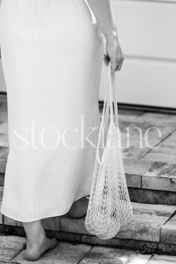 Vertical black and white stock photo of a barefoot woman in a white dress holding a bag with grapefruits.