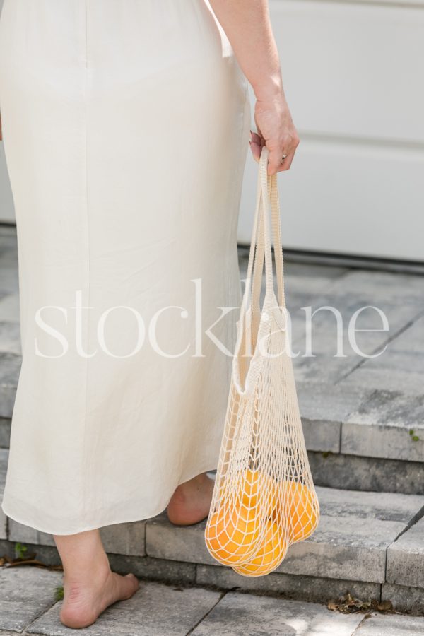 Vertical stock photo of a barefoot woman in a white dress holding a bag with grapefruits.