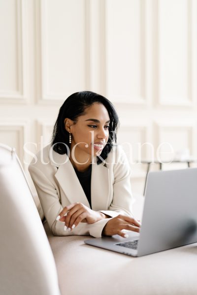 Vertical stock photo of a woman wearing a white suit, working on a laptop computer.