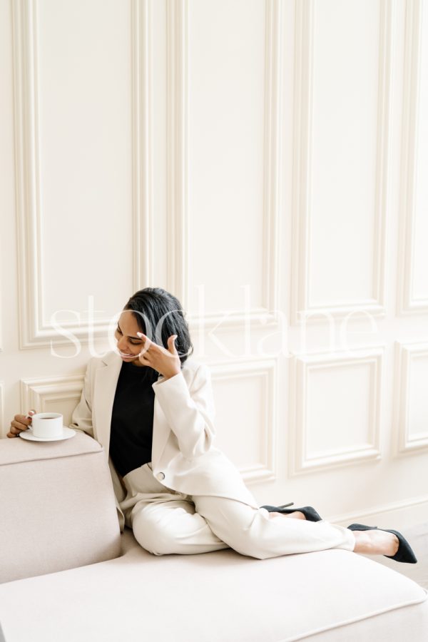 Vertical stock photo of a woman wearing a white suit, sitting on a blush pink couch, drinking a cup of coffee.