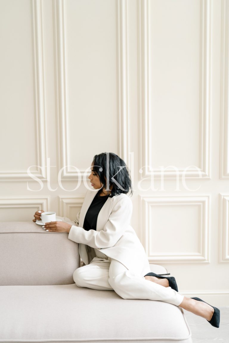 Vertical stock photo of a woman wearing a white suit, sitting on a blush pink couch, drinking a cup of coffee.