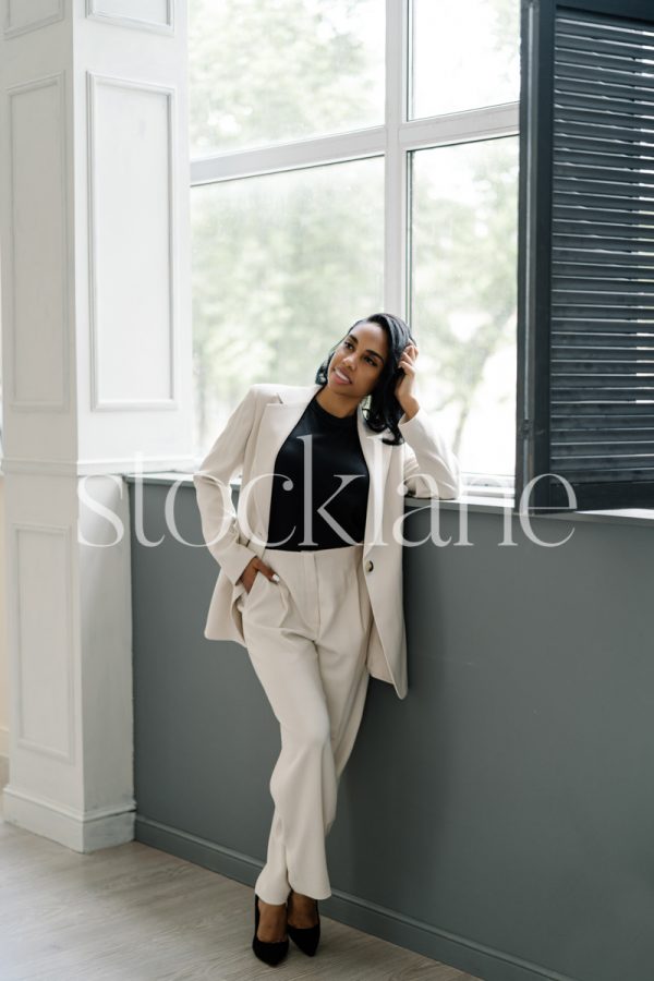 Vertical stock photo of a woman wearing a white suit leaning by a window.