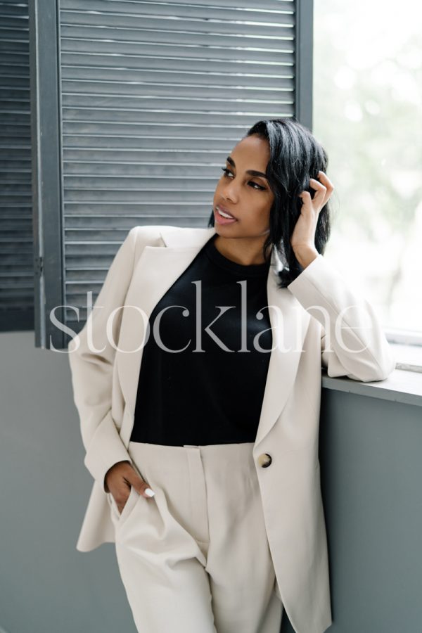 Vertical stock photo of a woman wearing a white suit leaning by a window.