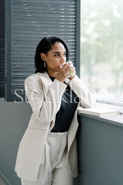 Vertical stock photo of a woman wearing a white suit drinking coffee by a window.