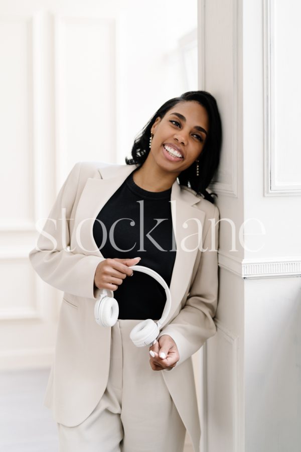 Vertical stock photo of a woman wearing a white suit, holding headphones, leaning against a pillar.