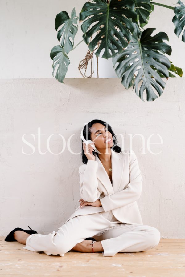 Vertical stock photo of a woman wearing a white suit, sitting on the floor, with headphones.