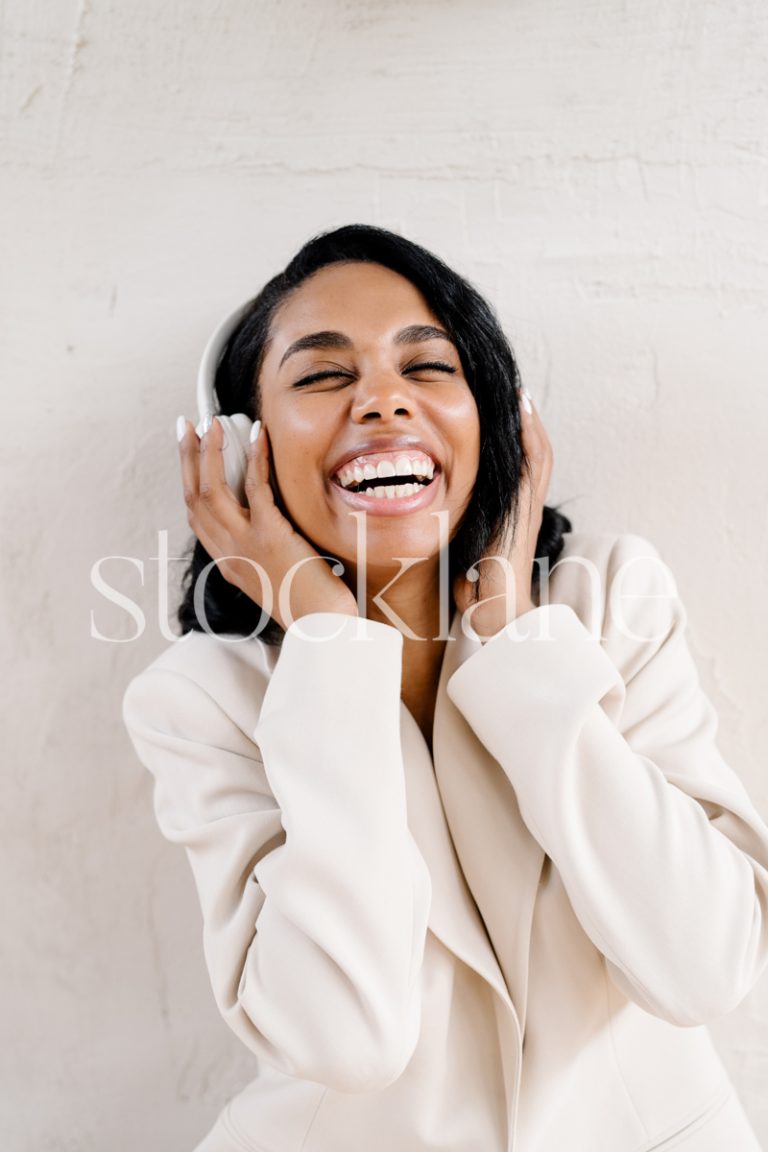 Vertical stock photo of a woman wearing a white suit, with headphones, smiling.