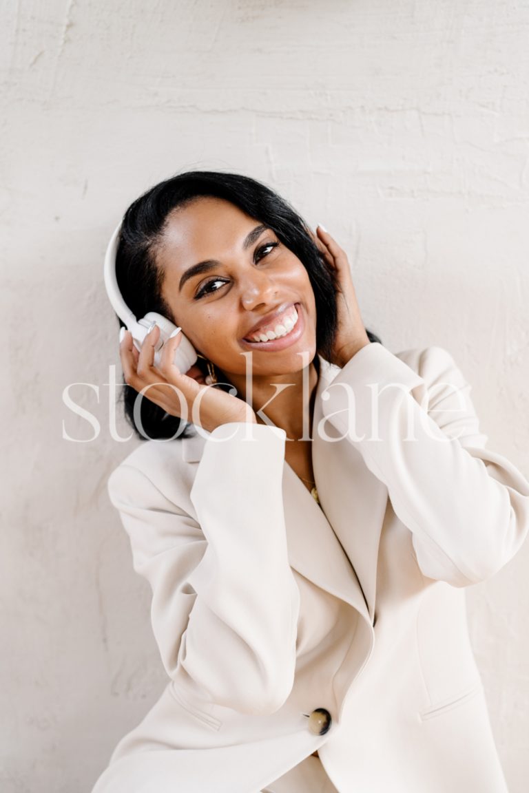 Vertical stock photo of a woman wearing a white suit, with headphones, smiling.