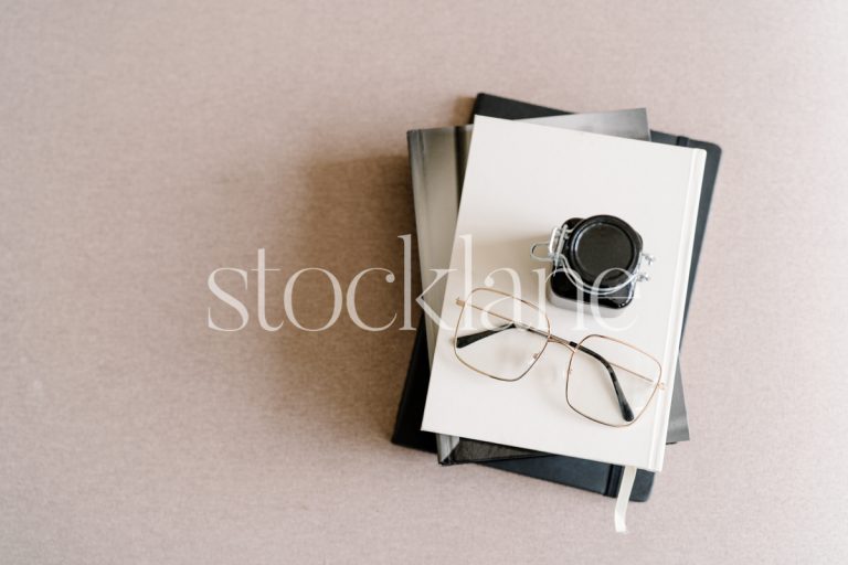 Horizontal stock photo of a pile of books with a pair of glasses and a bottle of ink, on a blush pink background.