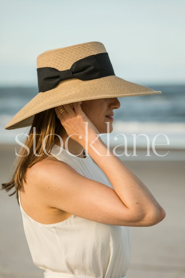 Vertical stock photo of a woman at the beach, wearing a white dress and a hat.