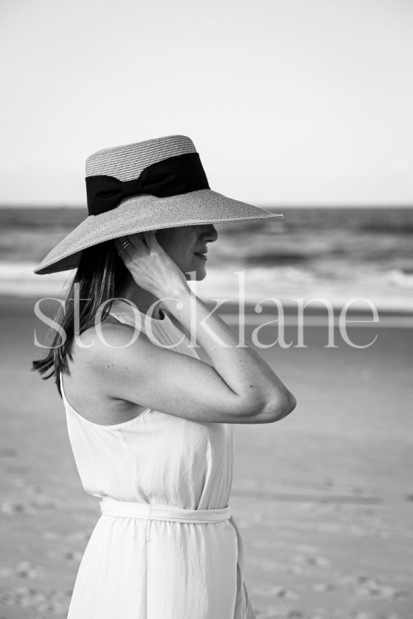 Vertical black and white stock photo of a woman at the beach, wearing a white dress and a hat.