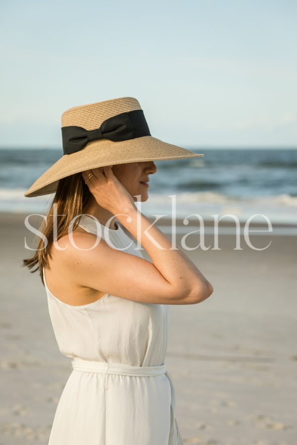 Vertical stock photo of a woman at the beach, wearing a white dress and a hat.