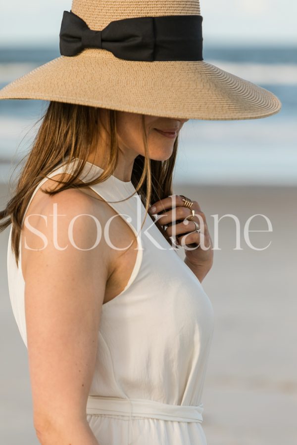 Vertical stock photo of a woman at the beach, wearing a white dress and a hat.