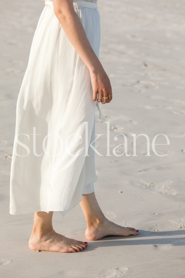 Vertical stock photo of a barefoot woman at the beach, wearing a white dress.