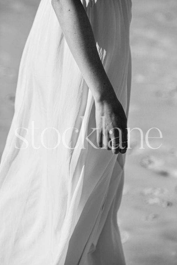 Vertical black and white stock photo of a barefoot woman at the beach, wearing a white dress.